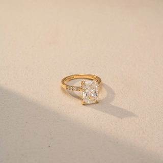 Side view of a radiant-cut diamond engagement ring in 14 KT Yellow Gold, featuring a radiant-cut lab-grown diamond with pavé-set diamonds along the band, displayed against a beige backdrop.