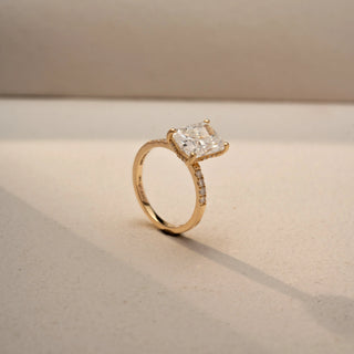 Angled view of a radiant-cut diamond engagement ring in 14 KT Yellow Gold, featuring a radiant-cut lab-grown diamond with pavé-set diamonds along the band, displayed against a beige backdrop.