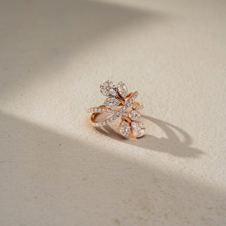 Side view of a floral crossover statement ring in 14 KT Rose Gold, featuring marquise and pear-shaped lab-grown diamonds arranged as blooming leaves on intertwined pavé-set diamond bands, displayed against a beige backdrop.