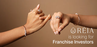 Two hands adorned with Reia Lab Grown diamond rings and sleek bracelets, gently reaching towards each other against a warm brown background, accompanied by “Franchise Investors” promotional branding for Reia.