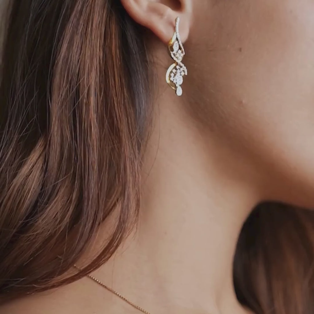  Dangling lab-grown diamond earrings by Réia in 14KT Yellow Gold, featuring vine-inspired curves and clustered diamonds, worn on the ear in soft natural light.