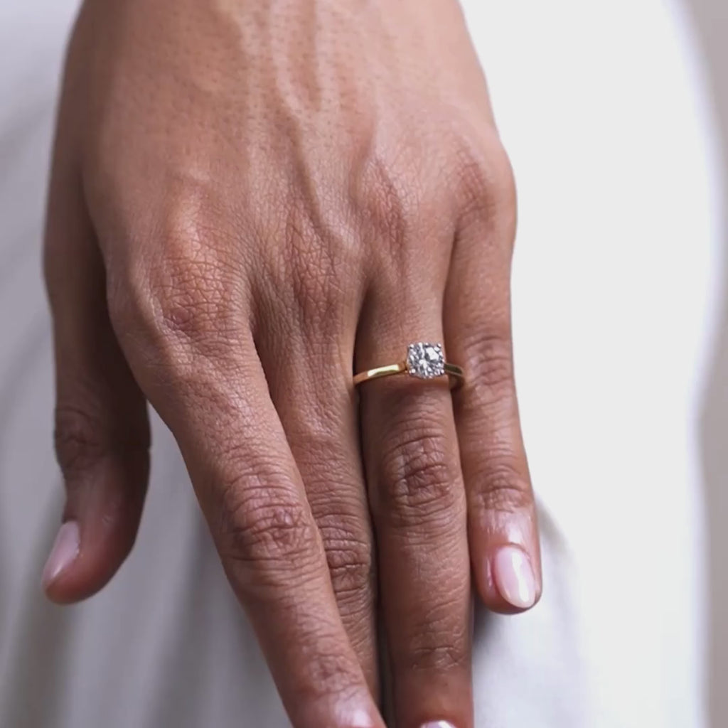 Styled on the hand, a lab-grown diamond solitaire ring with a princess-cut centre stone set on a slim yellow gold band, captured from a top-down lifestyle angle against a soft neutral background in natural light.